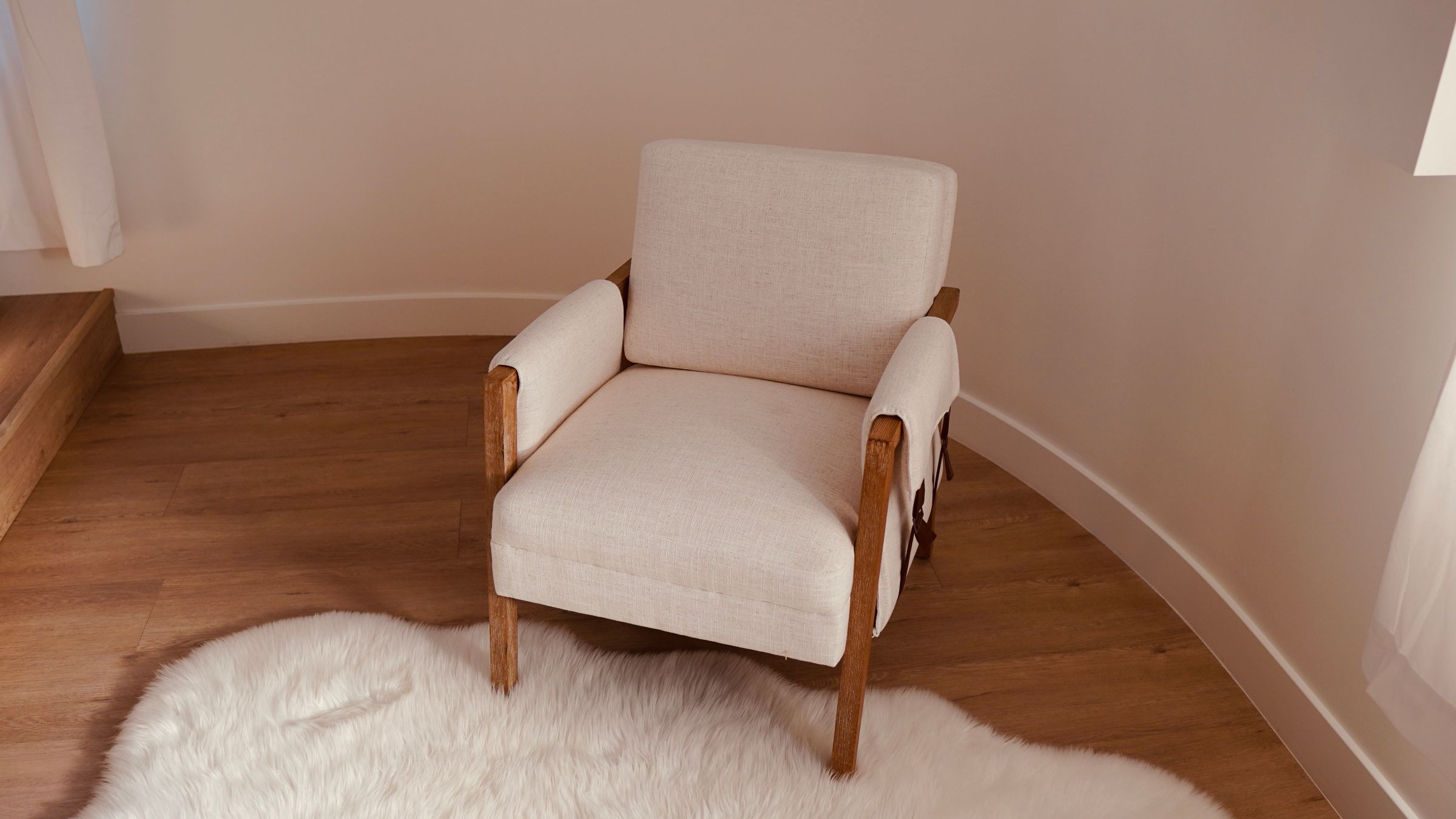 A cream upholstered armchair with wooden arms in the corner of a room, on an oak floor and a sheepskin rug.