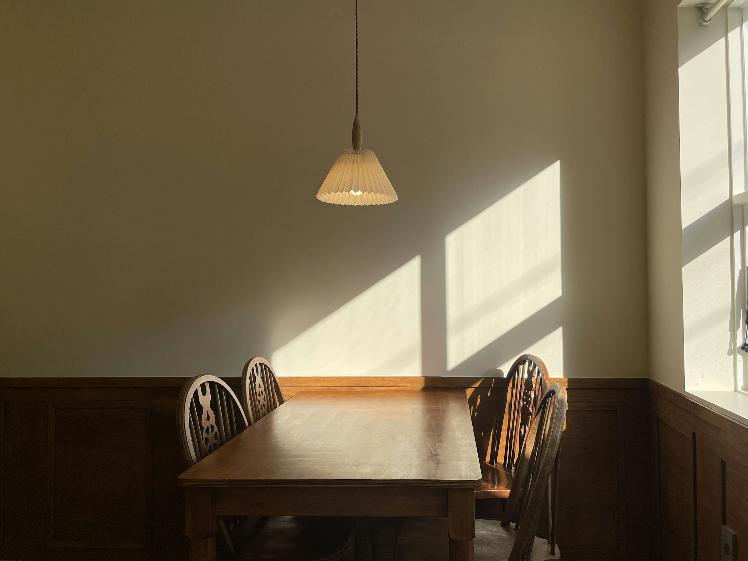 A wooden dining table and three chairs beside a window, a paper pendant lamp above, morning light across the wall.
