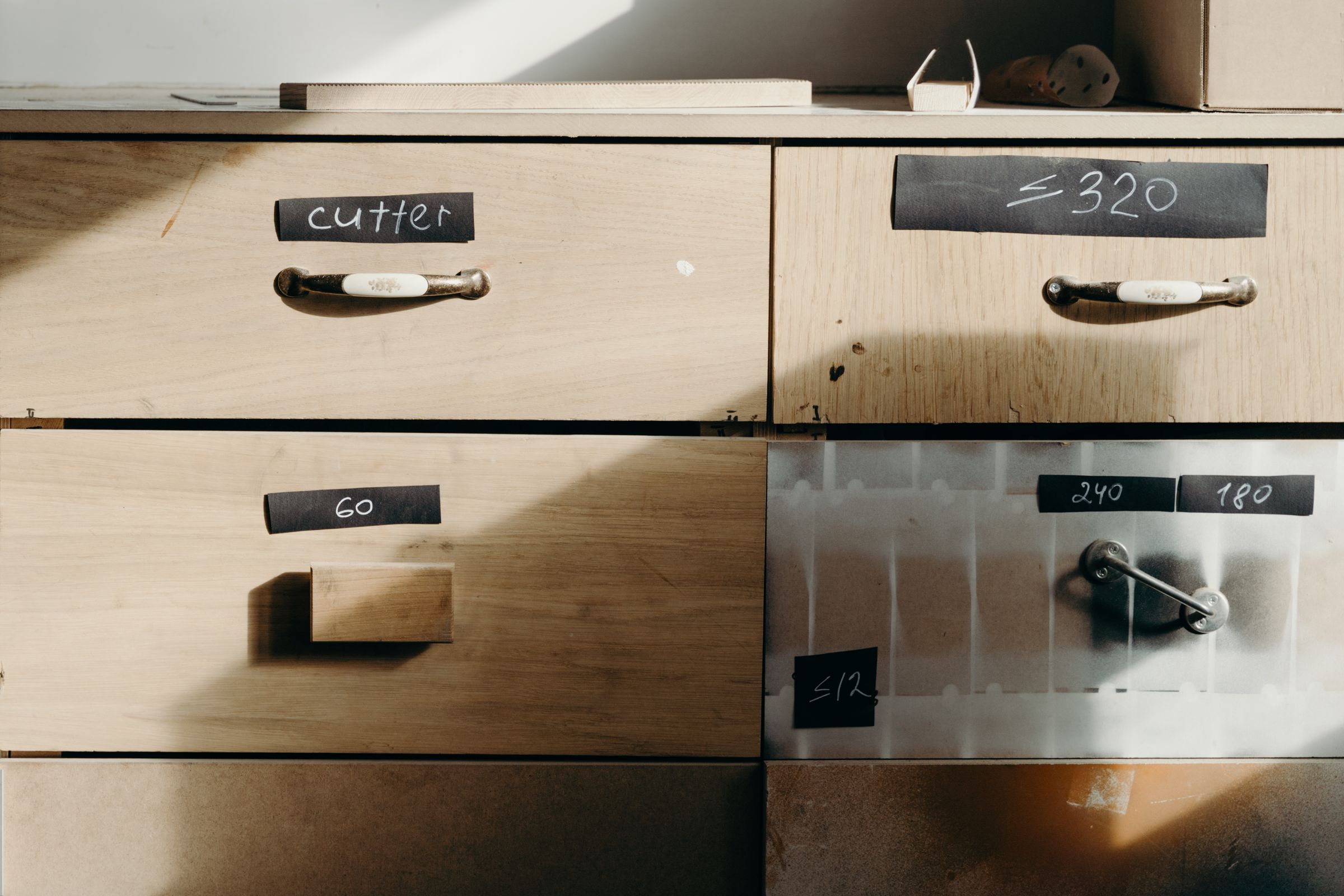 Pale wooden drawers with handwritten labels on black tape, lit by a window from the left.