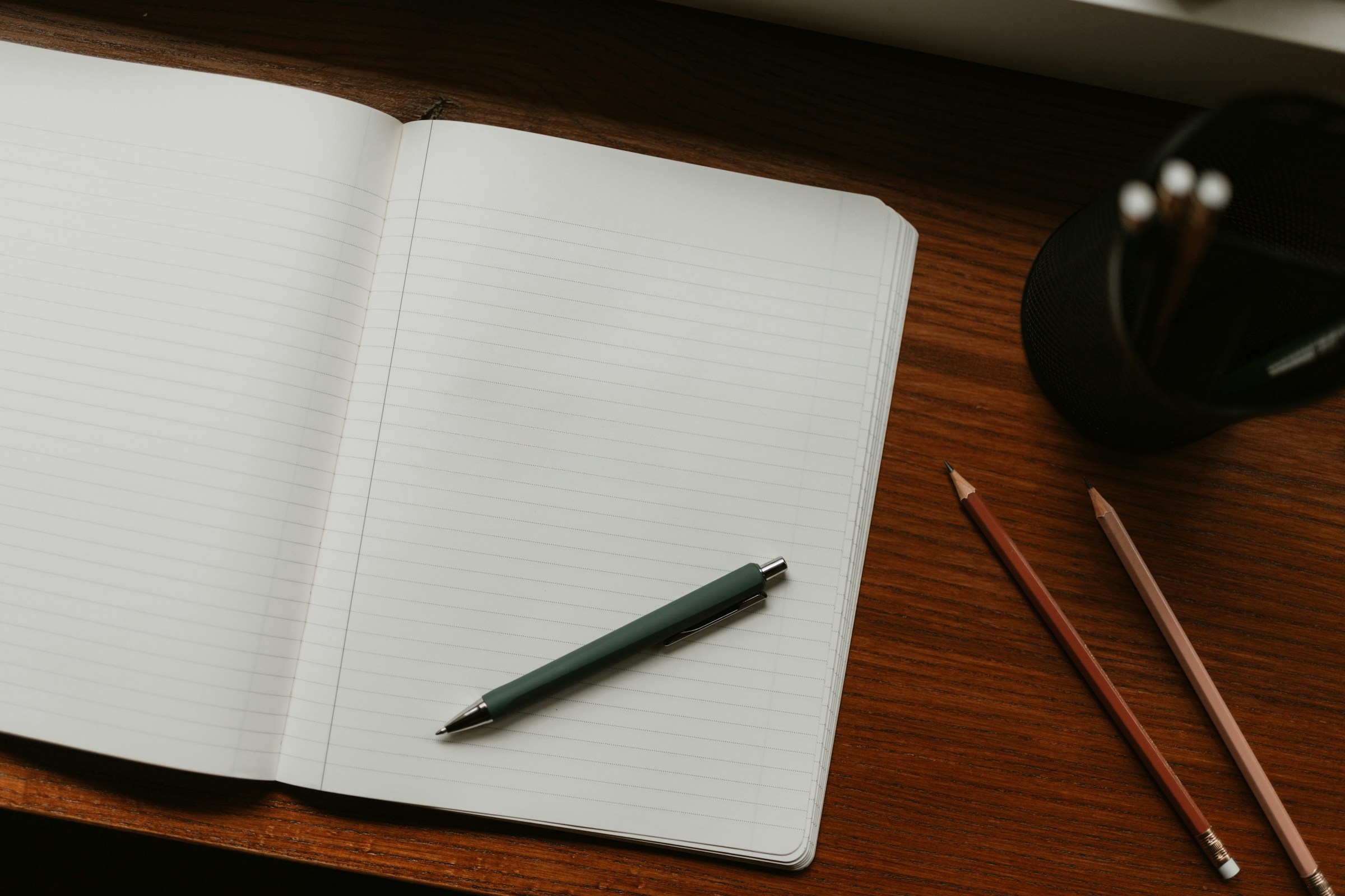 An open lined notebook with a pen laid on the blank page, two pencils and a cup of pencils on a dark wood desk.