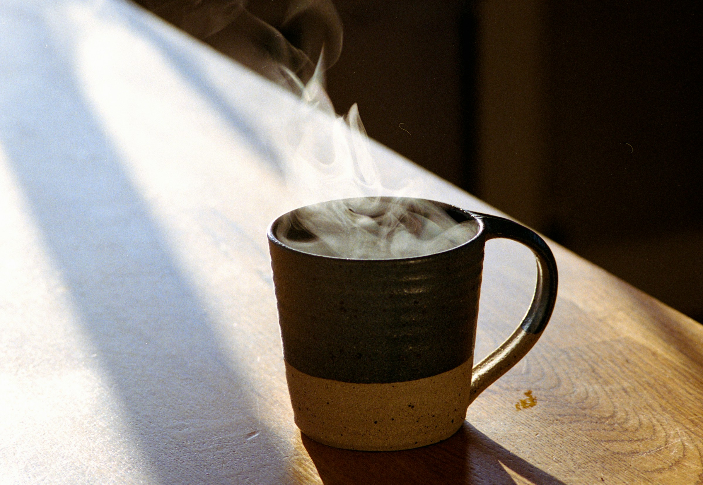 A stoneware mug with steam rising, on a wooden table in a band of morning sun.