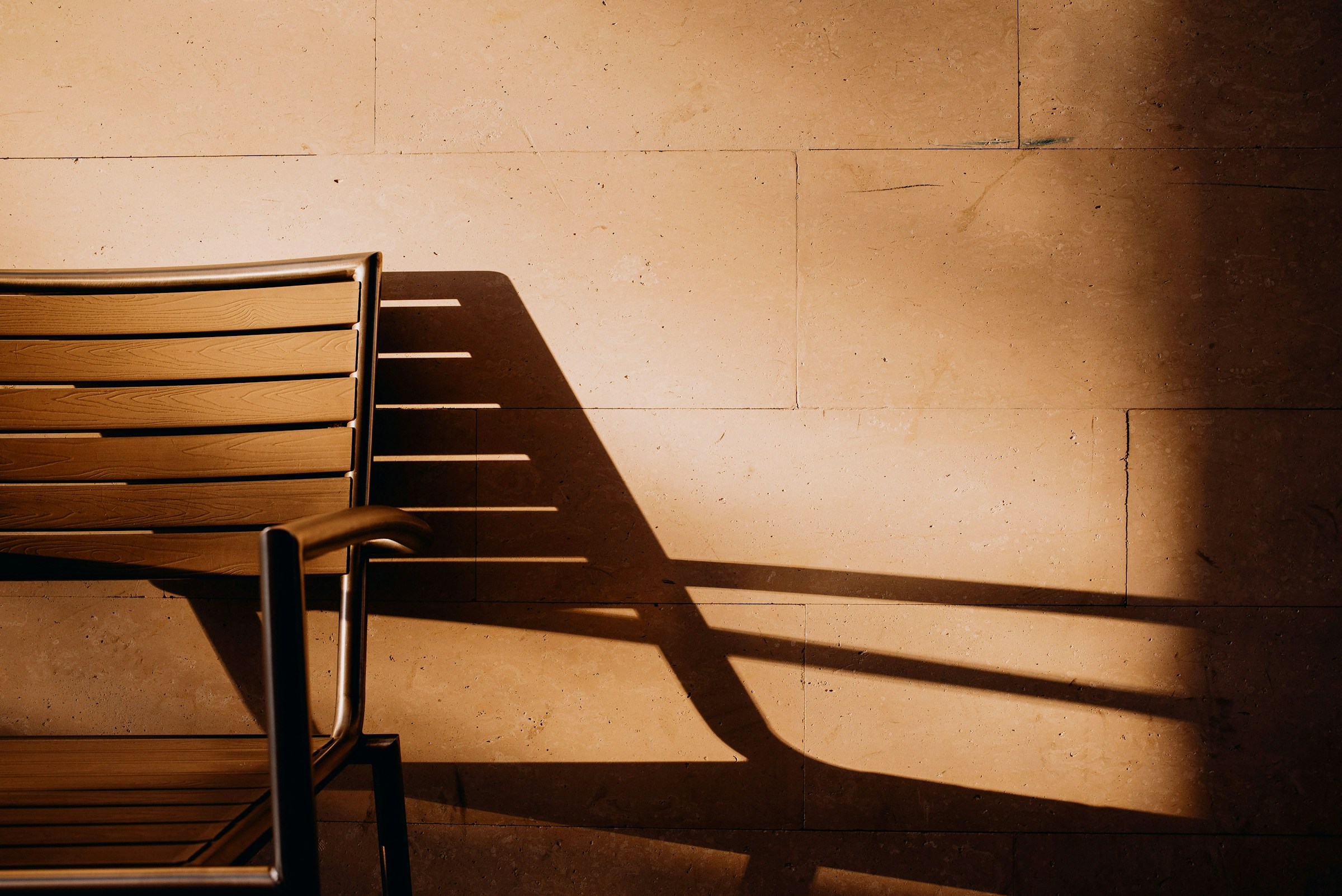 A wooden slat chair against a travertine wall, its shadow stretched by low sun.