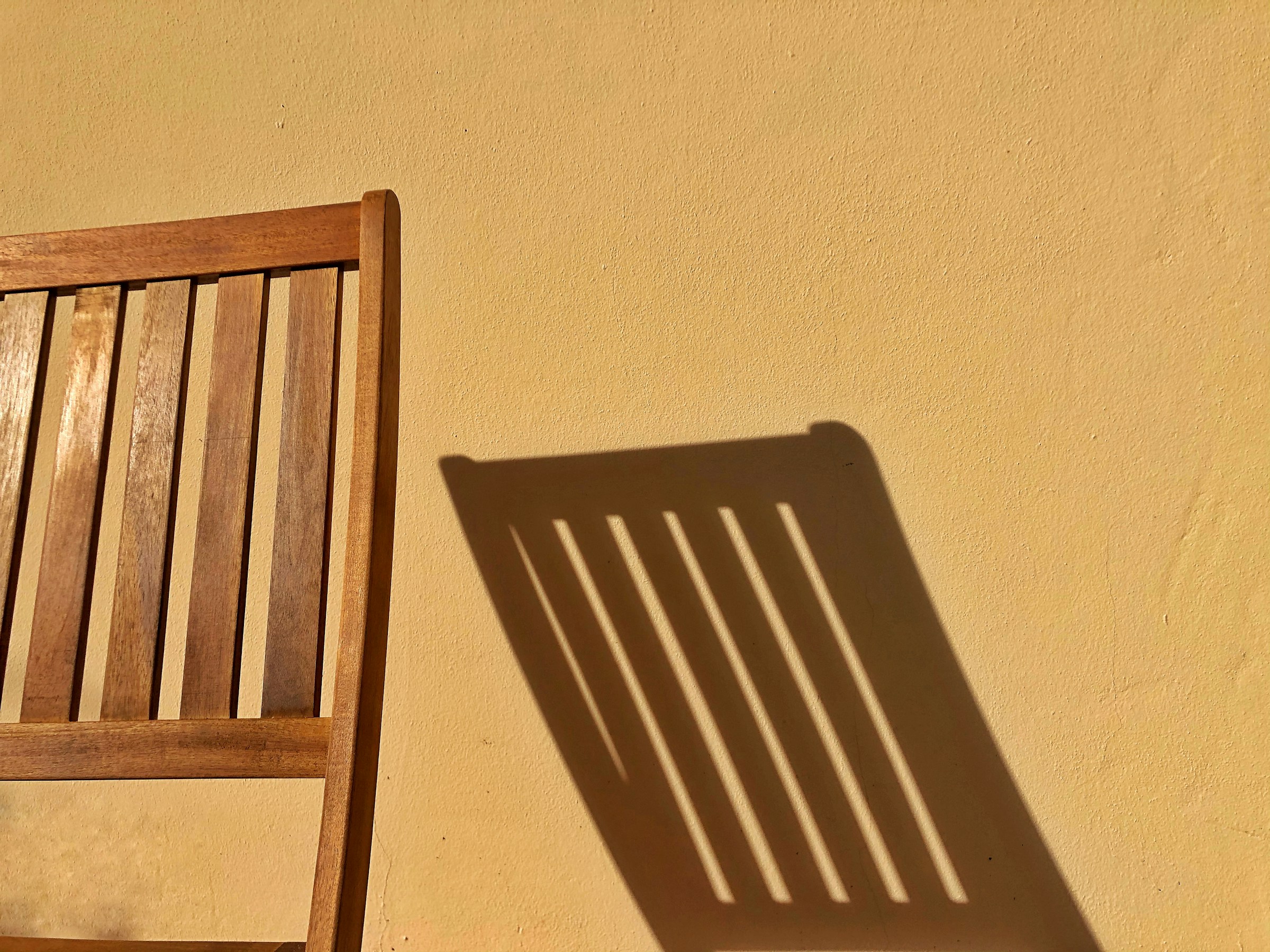 A wooden slat chair in afternoon sun, its shadow thrown across a plain warm wall.